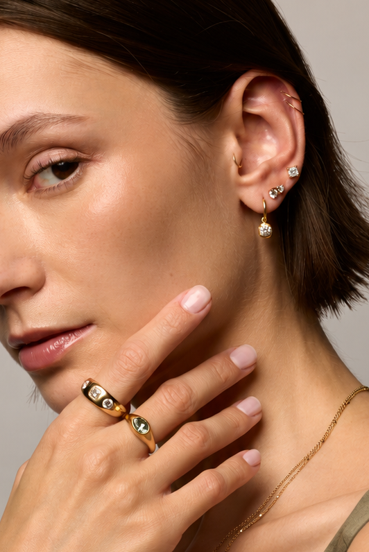 A close-up portrait of a woman with short brown hair wearing gold jewelry, including a gold ring with green and white stones on her finger and matching earrings on her ear, against a neutral gray background.