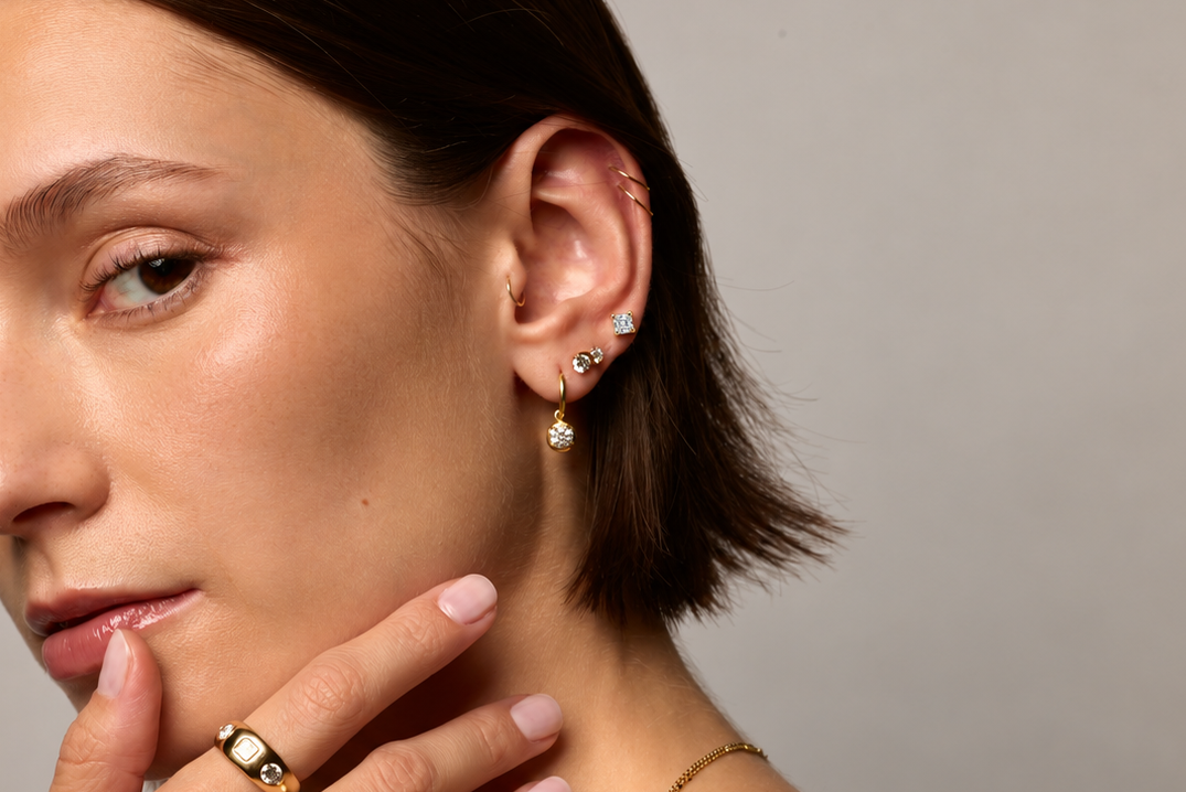 A close-up portrait of a woman with short brown hair wearing gold jewelry, including a ring with dark green stones on her finger and small hoop earrings, against a plain gray background.