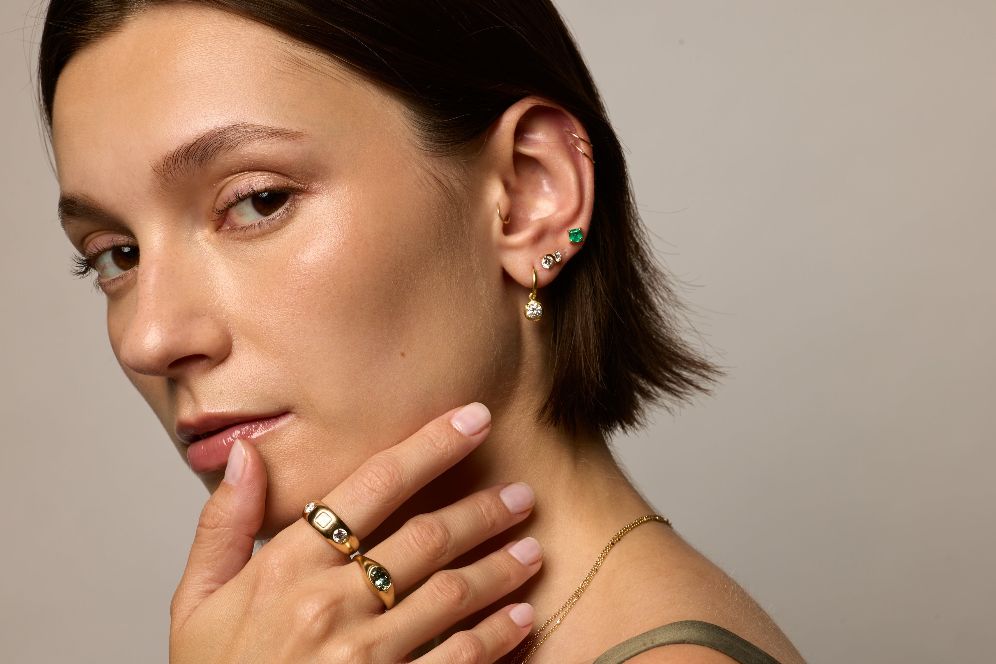 A close-up portrait of a woman with short dark hair wearing gold rings with green and white accents, along with small gold and green earrings, against a neutral light gray background.