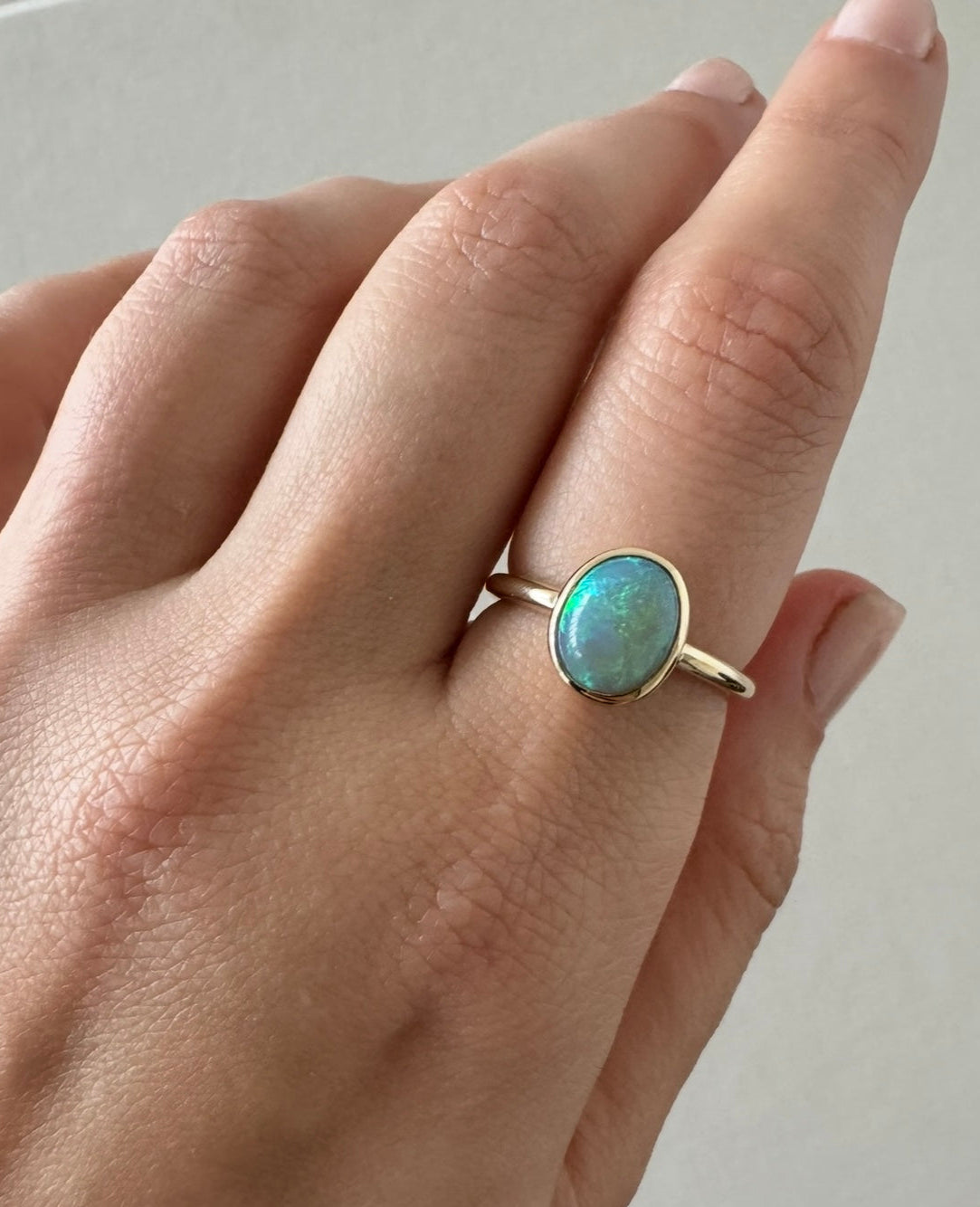 A gold ring with an oval green-blue opal gemstone worn on a person's finger, shown in a close-up shot.