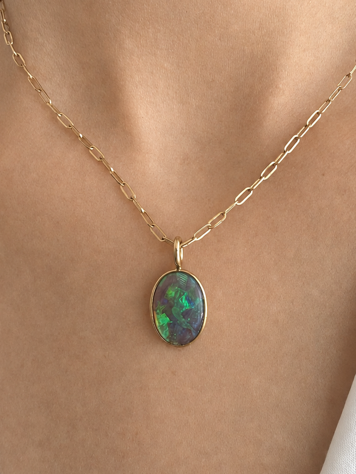 A gold chain necklace with an oval pendant featuring a green and blue iridescent opal, shown worn on a model's neck over a white collared shirt.