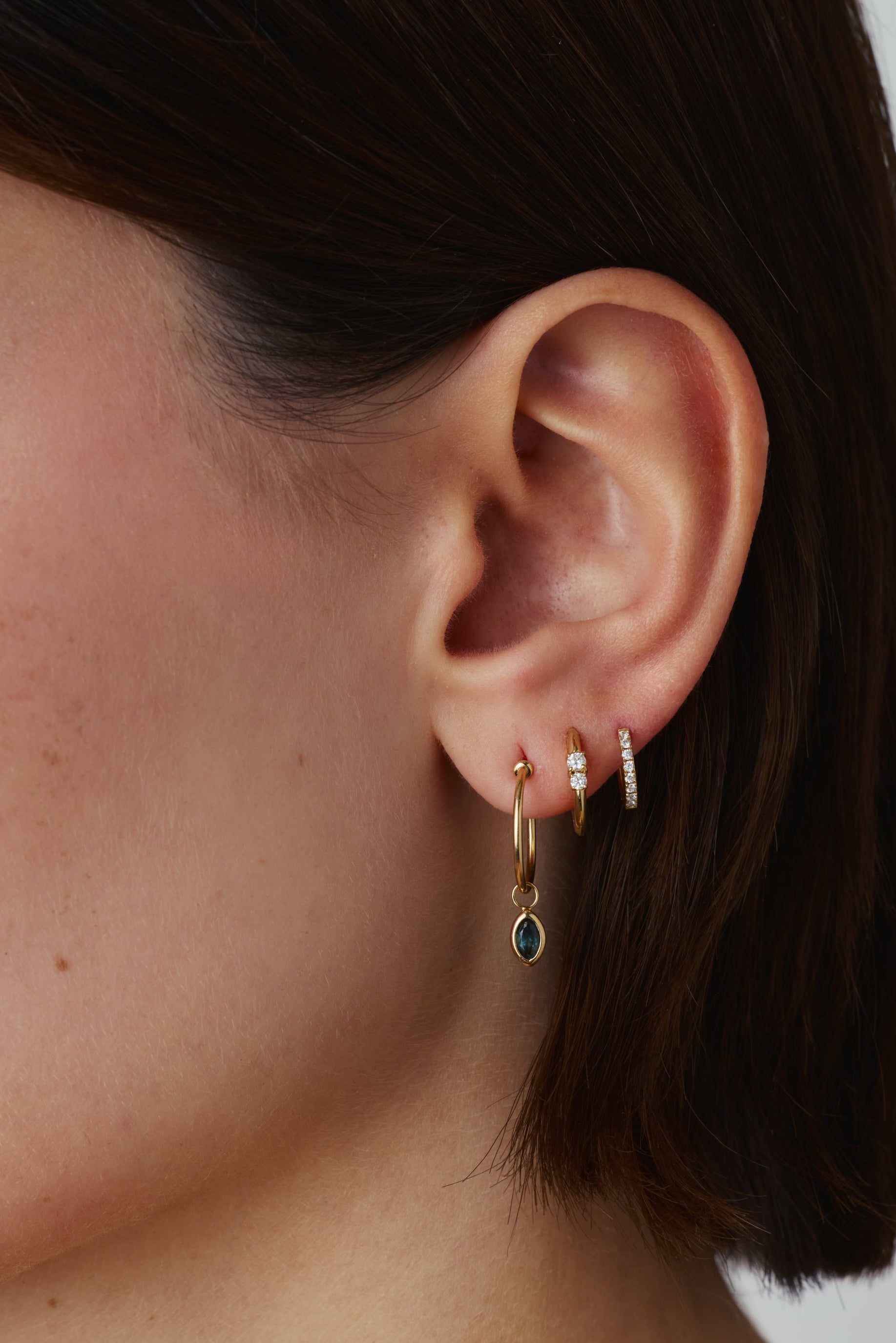 Close-up of a person's ear wearing multiple delicate earrings, including a small gold hoop with a dark green teardrop pendant and several thin gold huggie hoops, against a plain light gray background.
